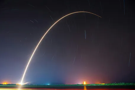 Long exposure photo of a rocket shooting into the night sky, forming a glowing arc over the horizon